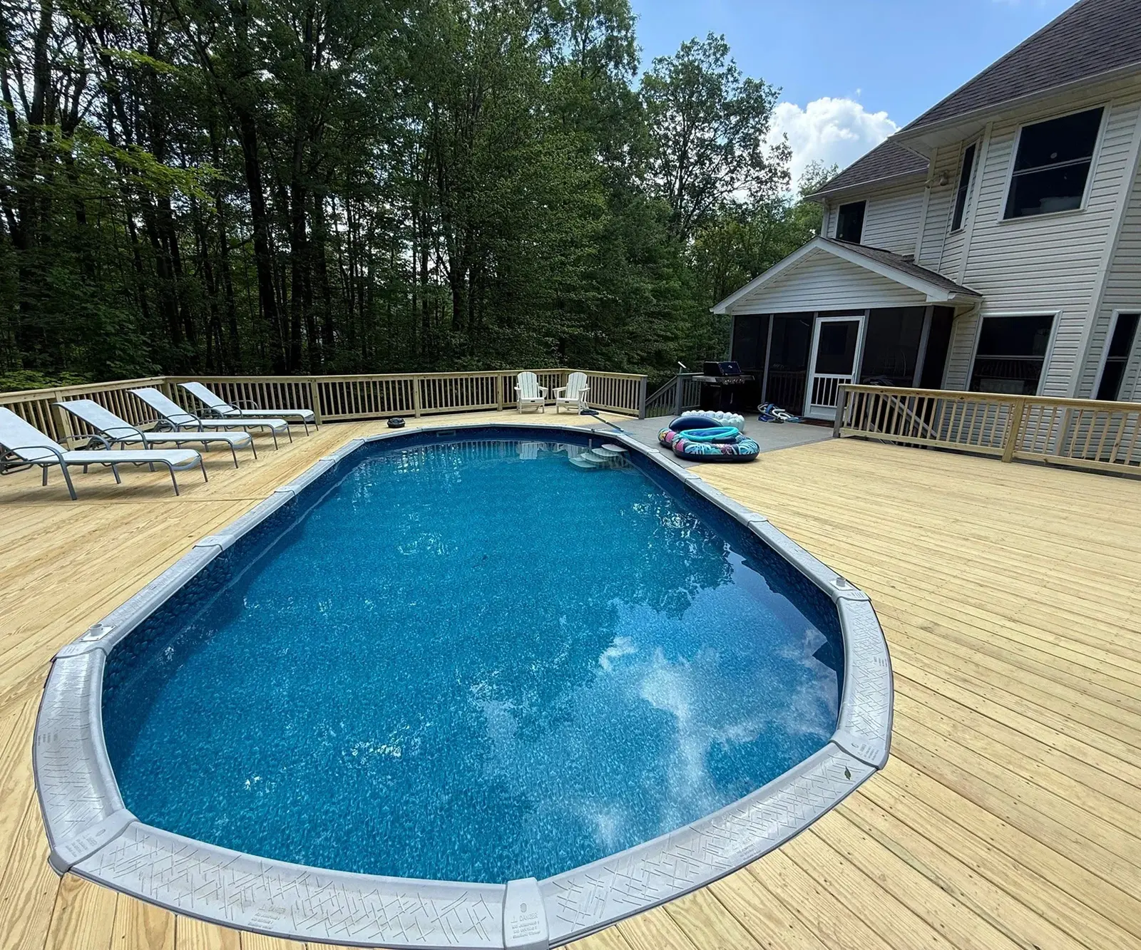 A backyard pool with a wooden deck and lounge chairs under a sunny sky.